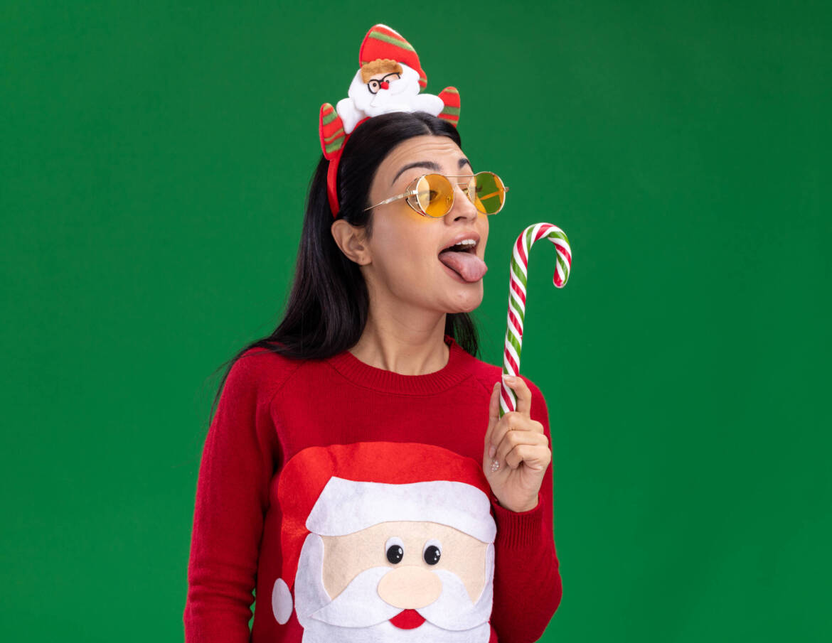 young-caucasian-girl-wearing-santa-claus-headband-sweater-with-glasses-holding-looking-traditional-christmas-candy-cane-showing-tongue-getting-ready-lick-it-isolated-green-wall-with-copy-space-scaled.jpg